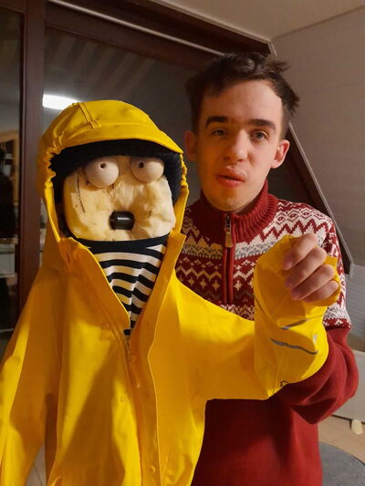 Auf dem Foto sieht man Raphael in einem Sweater mit Norwegermuster, der eine Puppe in gelber Regenjacke mit Kapuze hält.